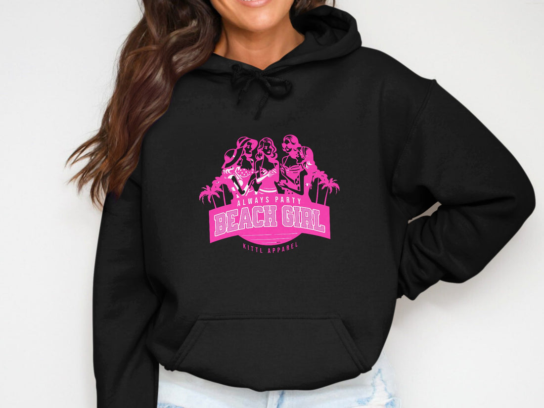 White hoodie from Garment Graphics features pink graphics of three cartoon women in beach attire with Always Party Beach Girl. Styled with a straw hat, sunglasses, sandals, and a camera on wood to complete the perfect casual look.