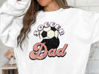 Someone is wearing a Garment Graphics sweatshirt showcasing Soccer Dad in pink, cursive font with stars. The retro design features a classic black and white soccer ball above Dad for nostalgia.