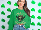 A woman in a Garment Graphics green sweater, showcasing a skull design and Liberty Motorcycles Specialist text, exudes biker style as she stands by a shamrock-adorned wall. Paired with denim shorts, she smiles confidently.
