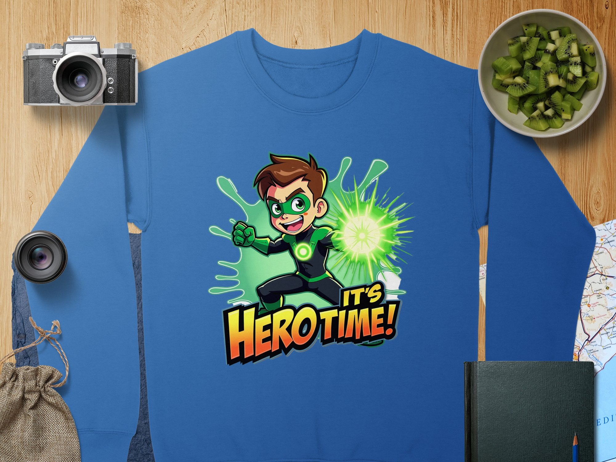 The Garment Graphics sweatshirt features a cartoon character with brown hair in a green suit, surrounded by green energy and the words Hero Time! Ideal for graphic print fans, it comes in unisex sizing. Accessories include a camera, lens, cucumber slices bowl, map, journal, and drawstring pouch.
