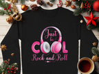 This Garment Graphics black sweater features Just be cool, rock and roll text with a pink headphones design. Surrounded by festive decorations like pine branches, ornaments, stars, and a cup of tea on a wooden surface—ideal for music enthusiasts and fashion lovers this Christmas season.
