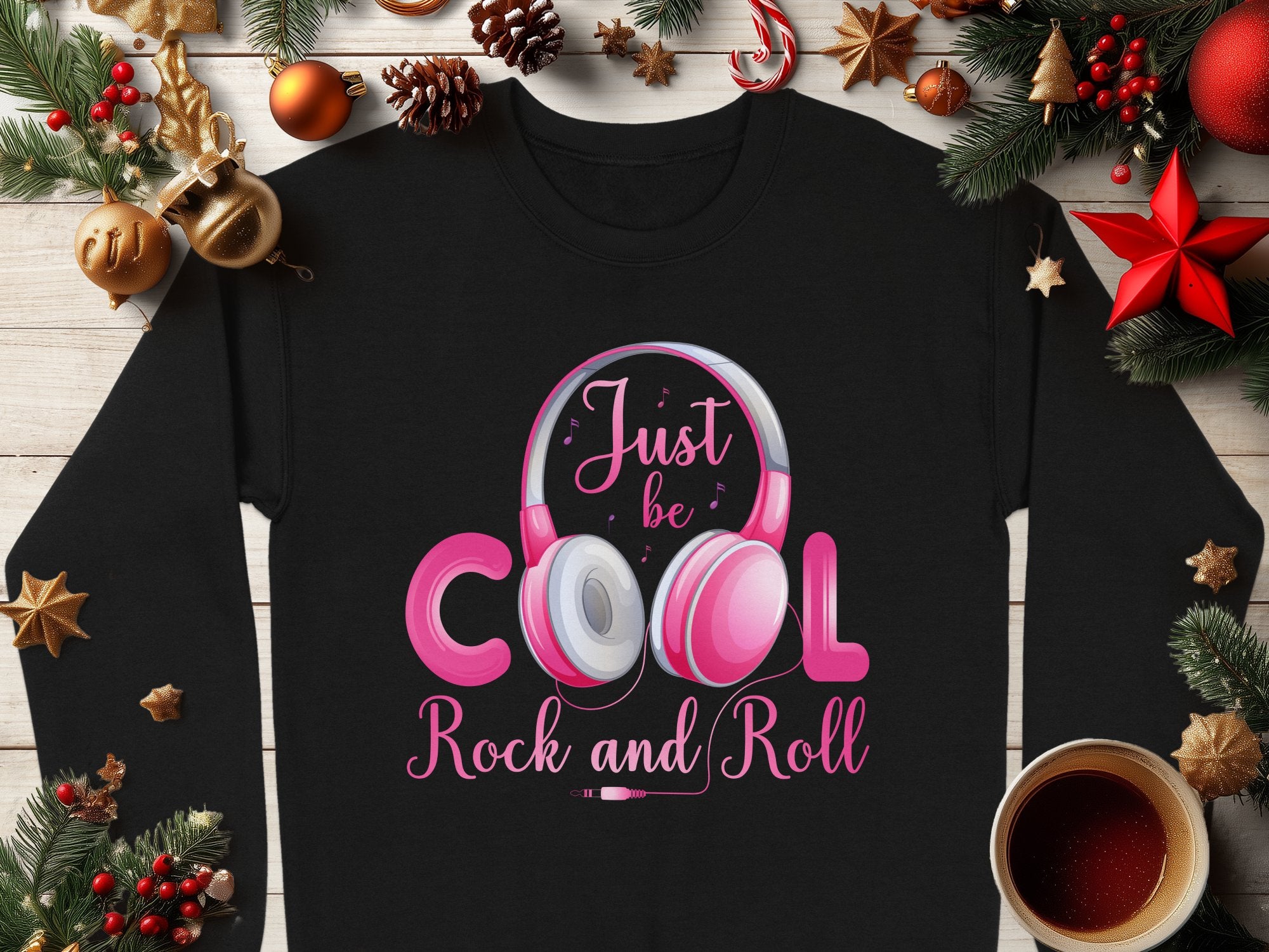 This Garment Graphics black sweater features Just be cool, rock and roll text with a pink headphones design. Surrounded by festive decorations like pine branches, ornaments, stars, and a cup of tea on a wooden surface—ideal for music enthusiasts and fashion lovers this Christmas season.