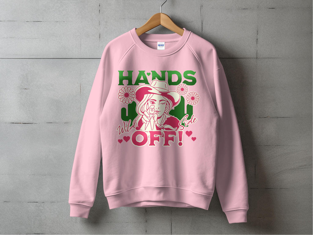 Garment Graphics Wild Side Sweatshirts feature a unisex pink sweatshirt with bold graphics on a wooden hanger against a gray wall, displaying HANDS OFF! in green and red, paired with an illustrated woman in a hat adorned with flowers and small red hearts.