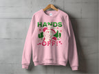 Garment Graphics Wild Side Sweatshirts feature a unisex pink sweatshirt with bold graphics on a wooden hanger against a gray wall, displaying HANDS OFF! in green and red, paired with an illustrated woman in a hat adorned with flowers and small red hearts.