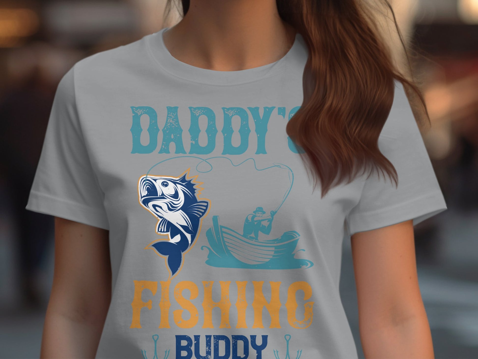 A person in a Garment Graphics gray womens fishing shirt featuring a large fish design, a person in a boat with a fishing rod, and the text Daddys Fishing Buddy in stylized letters.