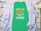 Garment Graphics presents a green T-shirt with Mine is so big I have to use both hands in bold white and orange letters, offering a humorous touch for fishing enthusiasts. Ideal under a light gray cardigan, its paired with white flowers and accessories for a playful angler-themed look.