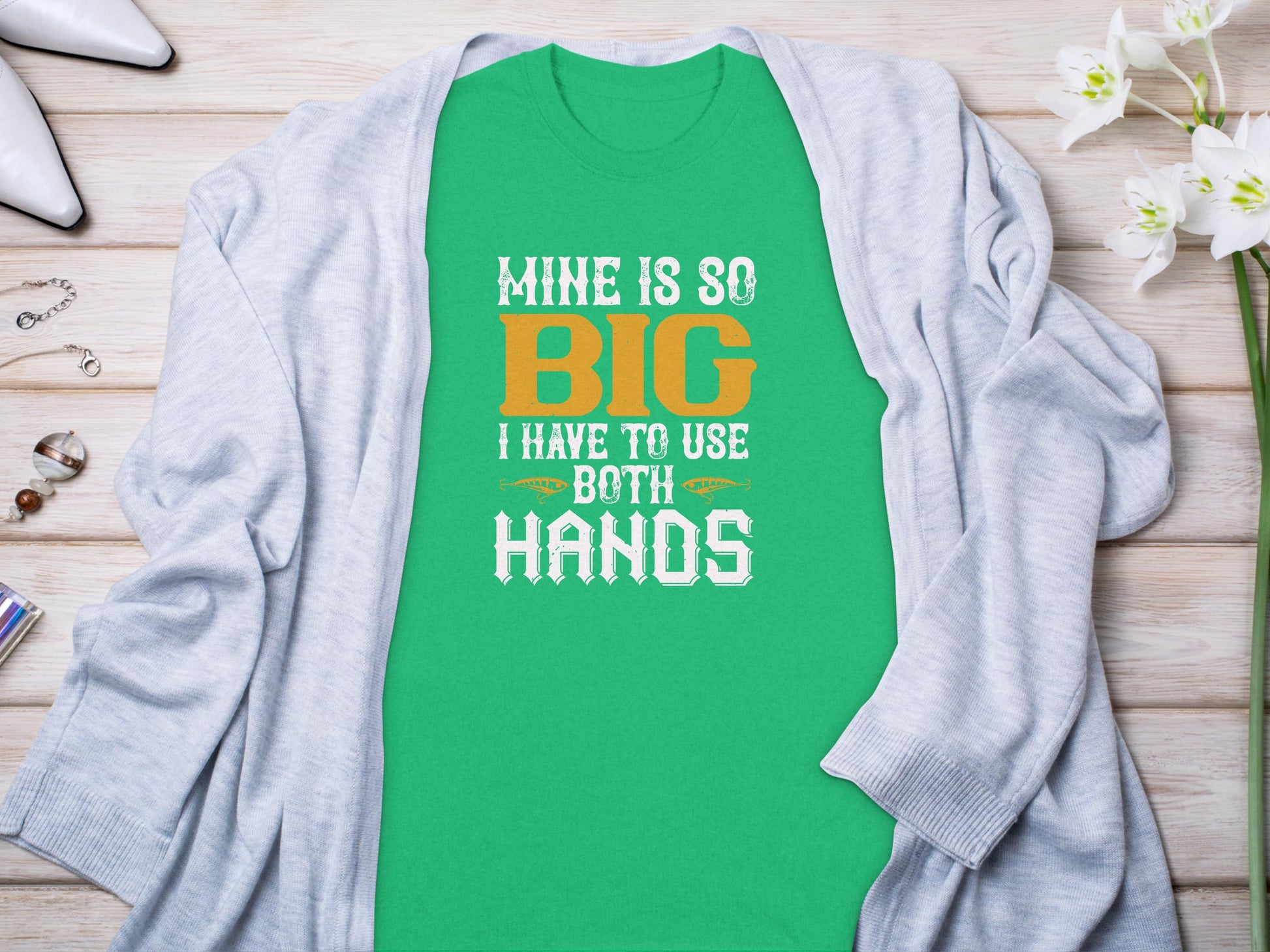 Garment Graphics presents a green T-shirt with Mine is so big I have to use both hands in bold white and orange letters, offering a humorous touch for fishing enthusiasts. Ideal under a light gray cardigan, its paired with white flowers and accessories for a playful angler-themed look.