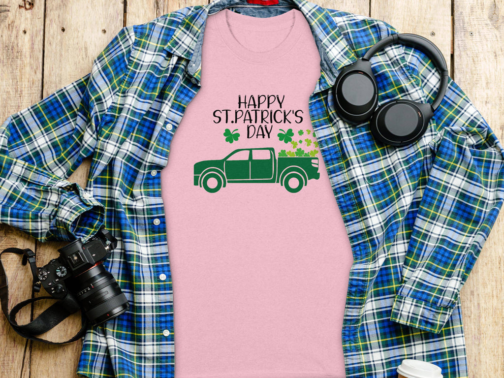 A flat lay showcases Garment Graphics pink T-shirt with an Irish pride twist, displaying a green truck and shamrock graphic reading Happy St. Patricks Day, set atop a blue plaid shirt. A camera, headphones, and coffee cup are nearby on the wooden surface.