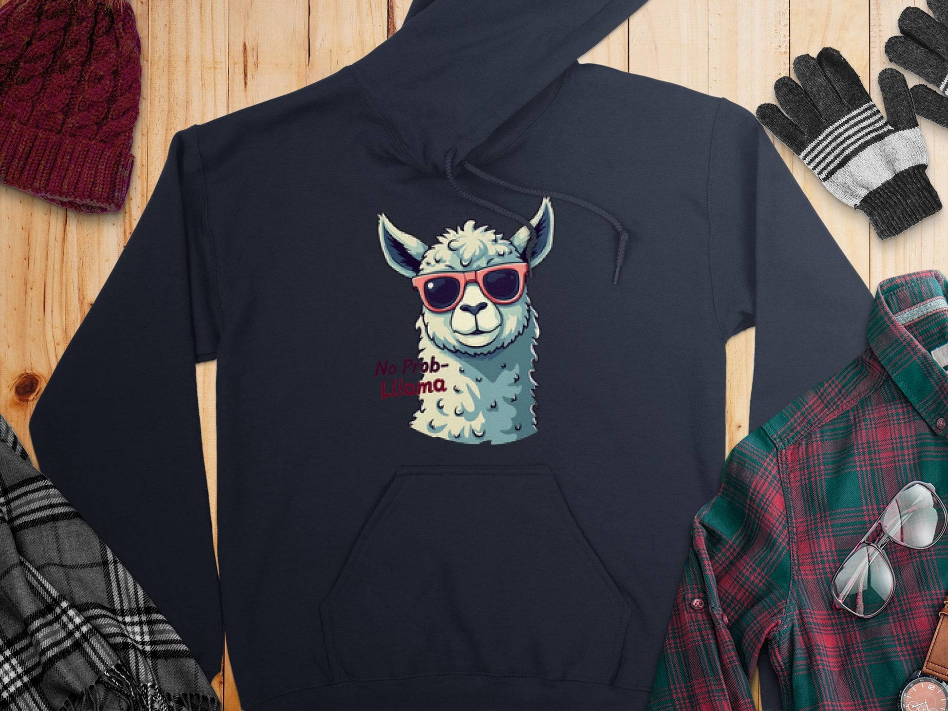 Garment Graphics offers a navy unisex hoodie with a cartoon llama in red sunglasses and No Prob-Llama text. Styled on wood, its paired with a red beanie, gloves, and plaid shirt.