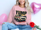 A person with long, wavy hair sits cross-legged, wearing a Garment Graphics pink sweatshirt with a rally car graphic. They hold a red rose next to metallic pink heart-shaped balloons and wear ripped blue jeans.