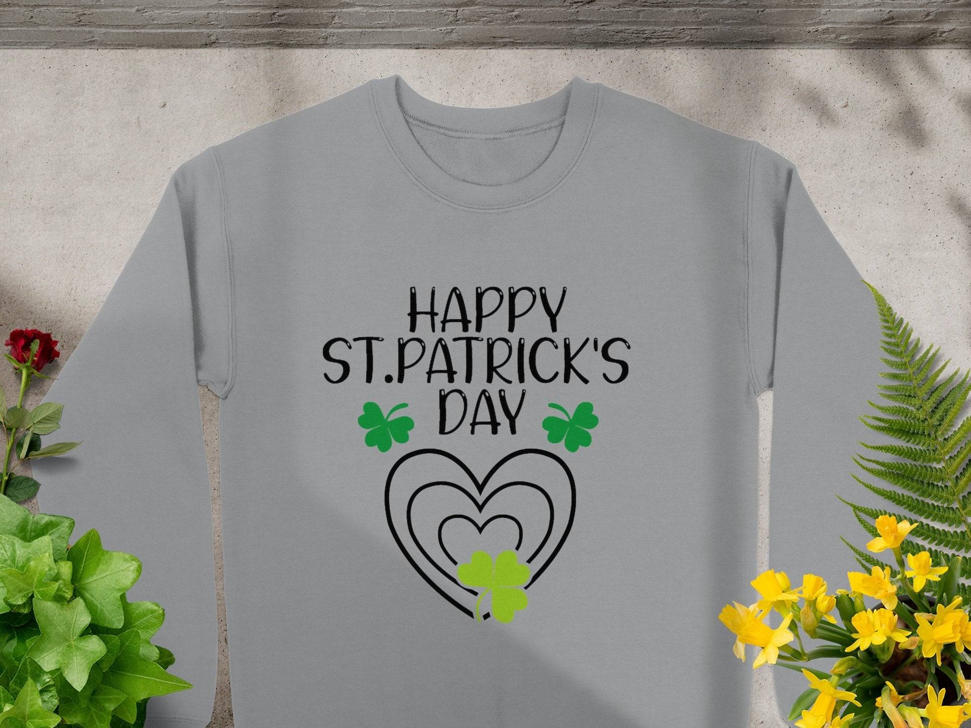 A Garment Graphics festive sweatshirt, featuring Happy St. Patricks Day in bold black on a gray base with a lively clover and green shamrocks, rests on concrete amidst vibrant green and yellow plants.