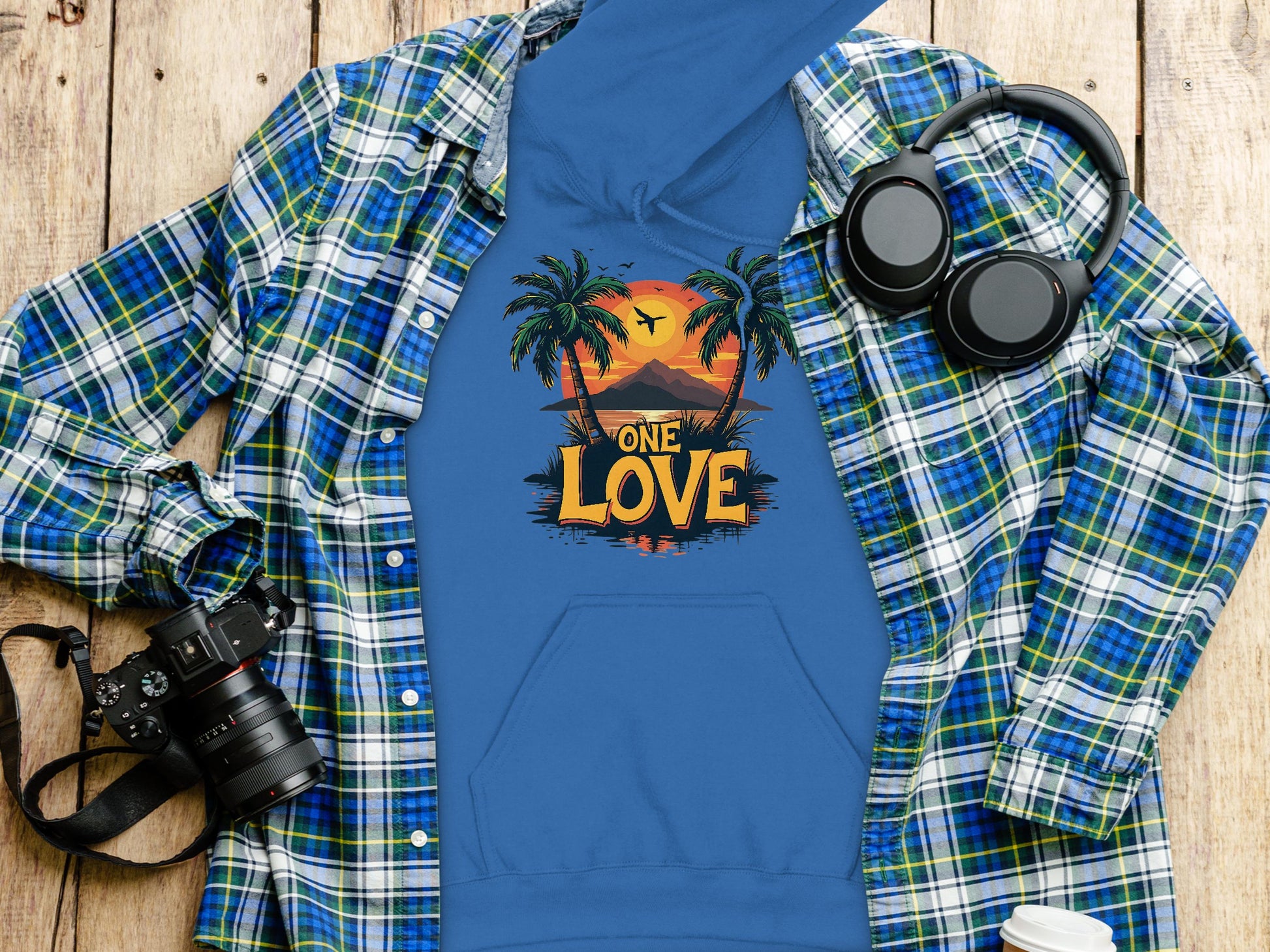 A Garment Graphics blue graphic hoodie with a tropical sunset, palm trees, and One Love text is laid on a plaid shirt. Nearby on the wooden surface are headphones, a camera, and a white cup lid, all showcasing casual elegance.