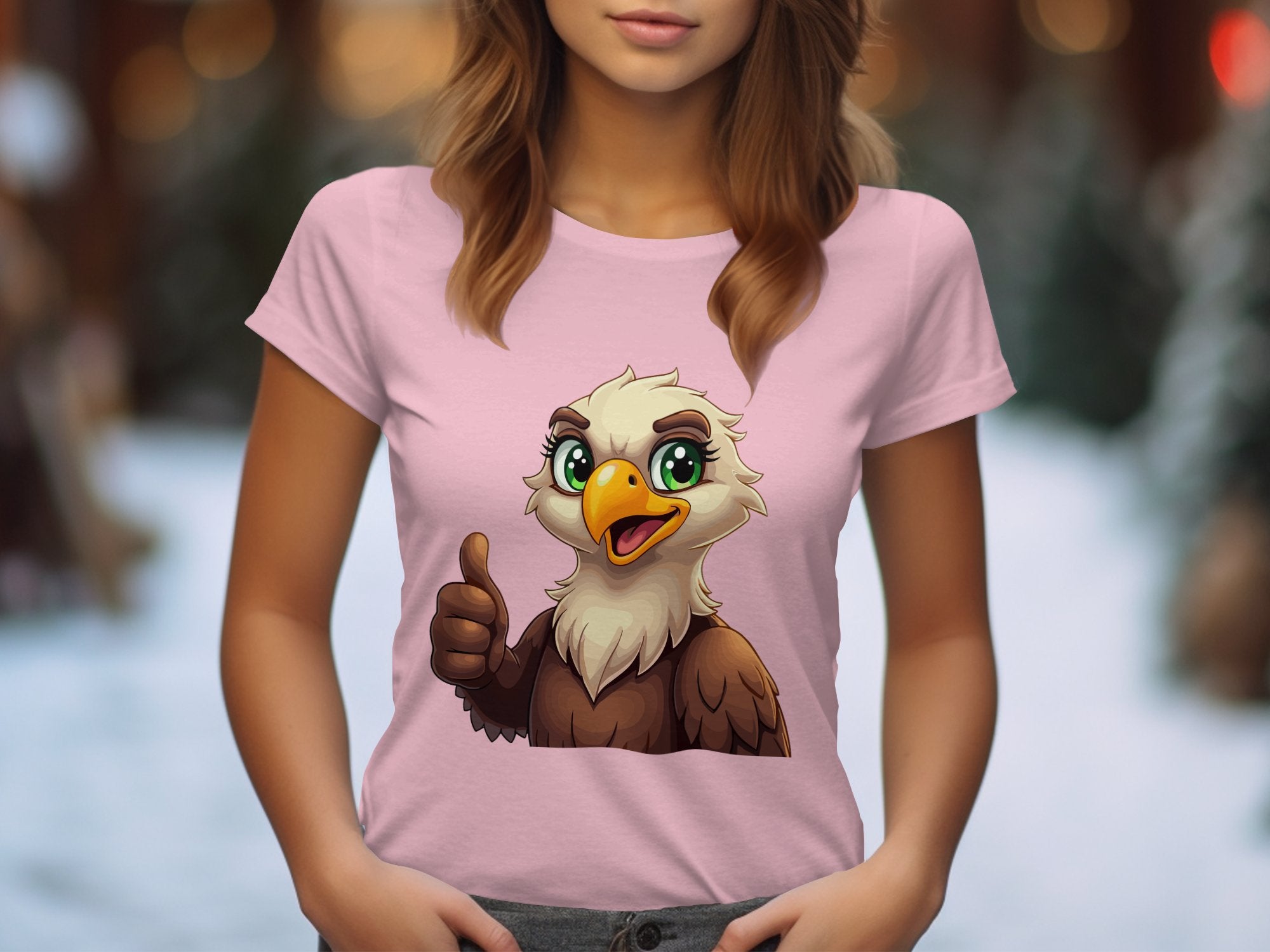 A person is wearing a Garment Graphics light pink cartoon T-shirt featuring an eagle with large, expressive eyes giving a thumbs-up. Ideal for animal lovers, its blurred natural color background suggests an outdoor setting.