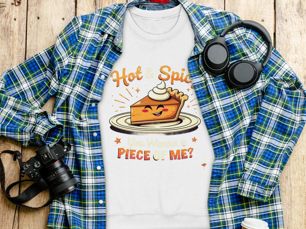 A Garment Graphics flat lay features a plaid shirt, a playful pie character T-shirt with Hot & Spicy, You Wanna a Piece of Me? text, unisex sweatshirts, alongside headphones, a camera, and a coffee cup on the wooden surface.