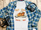 A Garment Graphics flat lay features a plaid shirt, a playful pie character T-shirt with Hot & Spicy, You Wanna a Piece of Me? text, unisex sweatshirts, alongside headphones, a camera, and a coffee cup on the wooden surface.