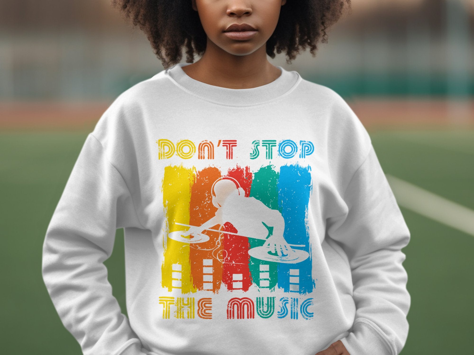 Wearing a Garment Graphics white sweatshirt with a colorful DJ design and turntables amid red, yellow, and blue hues, the person stands outdoors. The bold text reads Dont Stop the Music, making it perfect for music lovers while the blurred setting adds vibrant allure.