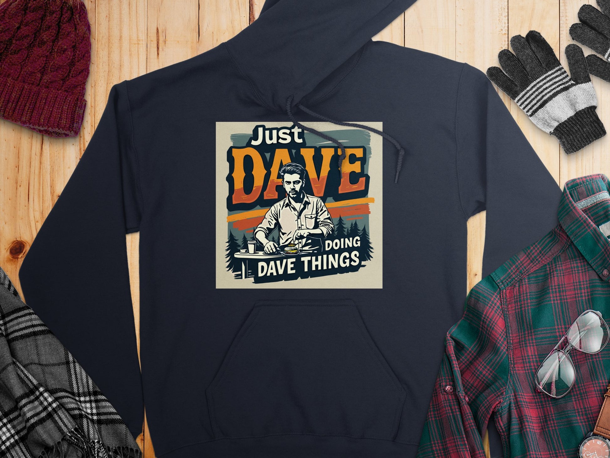 Garment Graphics navy graphic hoodie with Just Dave Doing Dave Things is crafted from medium-heavy fabric, ideal for cooler days. Its displayed on wood alongside a red beanie, checkered scarf, striped gloves, a plaid shirt, and glasses.