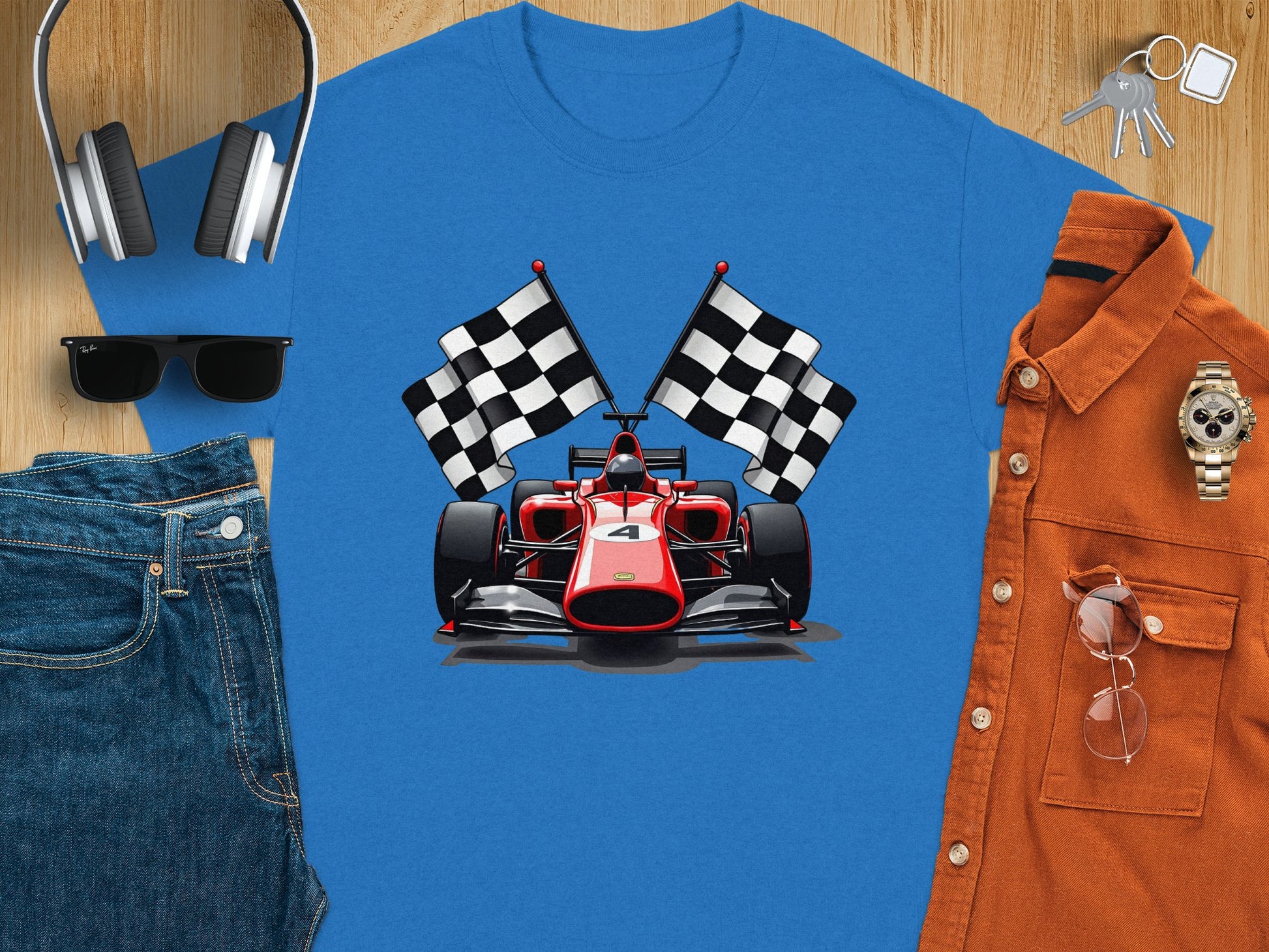 Garment Graphics blue T-shirt boasts a vivid red racing car graphic with two checkered flags, complemented by headphones, sunglasses, blue jeans, a brown button-up shirt, watch, keys, and smartphone on a light wooden surface. Ideal for any motorsport enthusiasts casual style.