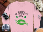 Garment Graphics pink sweater features Happy St. Patricks Day in black text with two green shamrocks and a clover kiss graphic. Its placed on a wooden surface with accessories like a leather case, camera, binoculars, headphones, belt, and rope. Celebrate the festivity with this distinct piece!.