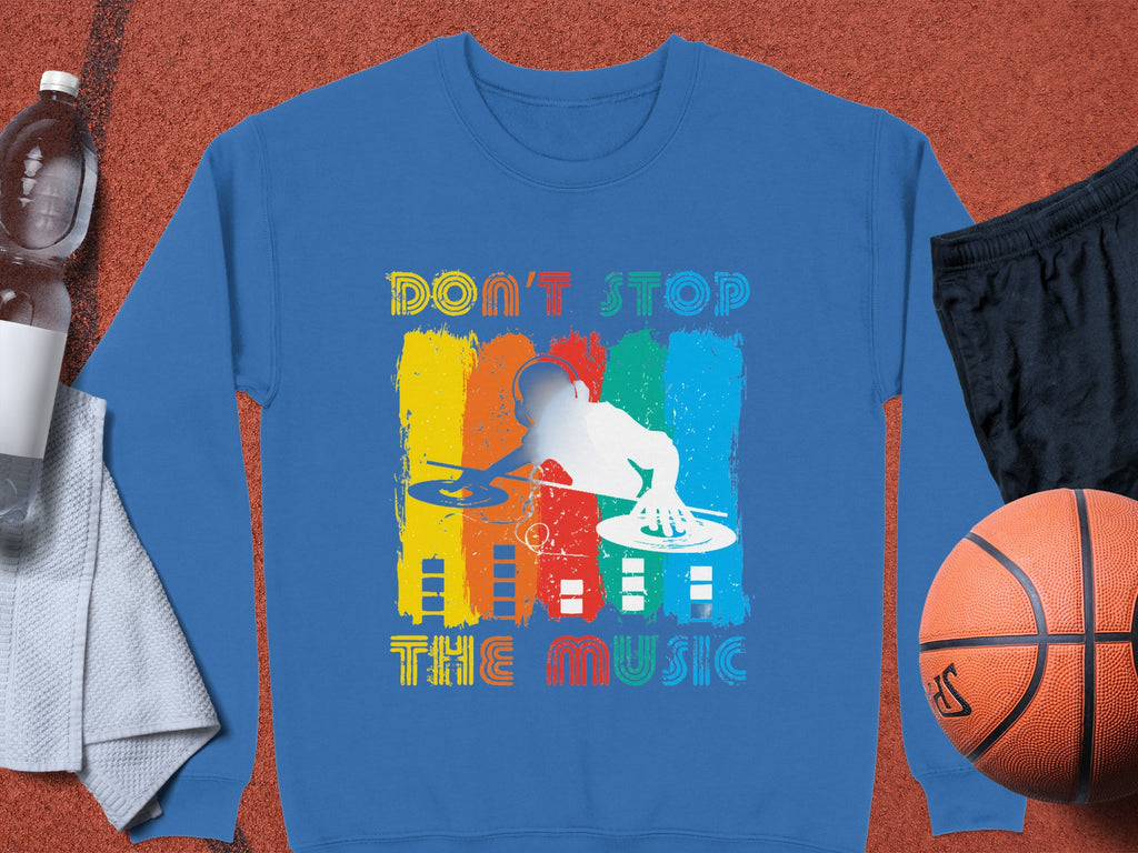 Garment Graphics offers a blue unisex DJ sweatshirt featuring a silhouette design on colorful stripes and the text Dont Stop the Music. Ideal for music enthusiasts, it is displayed with a basketball, black shorts, a towel, and a water bottle on a brown surface.