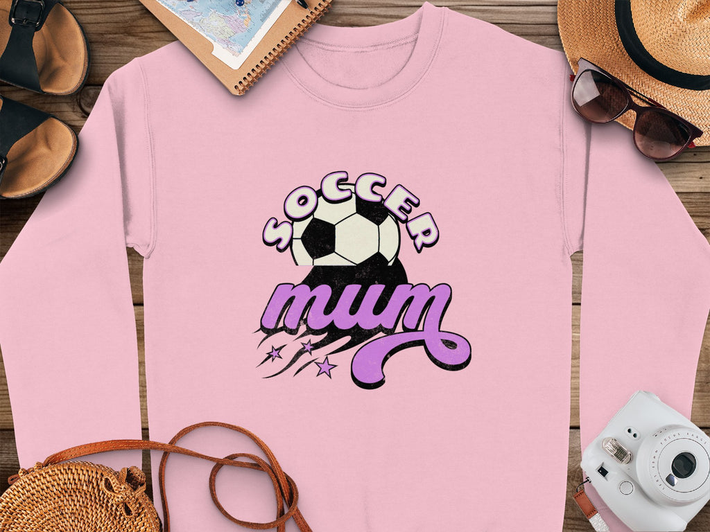 A Garment Graphics pink sweatshirt with a vintage Soccer Mum print and soccer ball graphic rests on a wooden surface with sandals, a notebook, stylish hat, sunglasses, and camera.