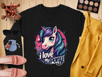 A unicorn enthusiasts perfect tee from Garment Graphics, this black shirt features a vibrant unicorn with colorful hair and horn, surrounded by stars. The text reads I love my pony. Styled on wood with makeup, sunglasses, a yellow jacket, pink shoes, and a small bag.