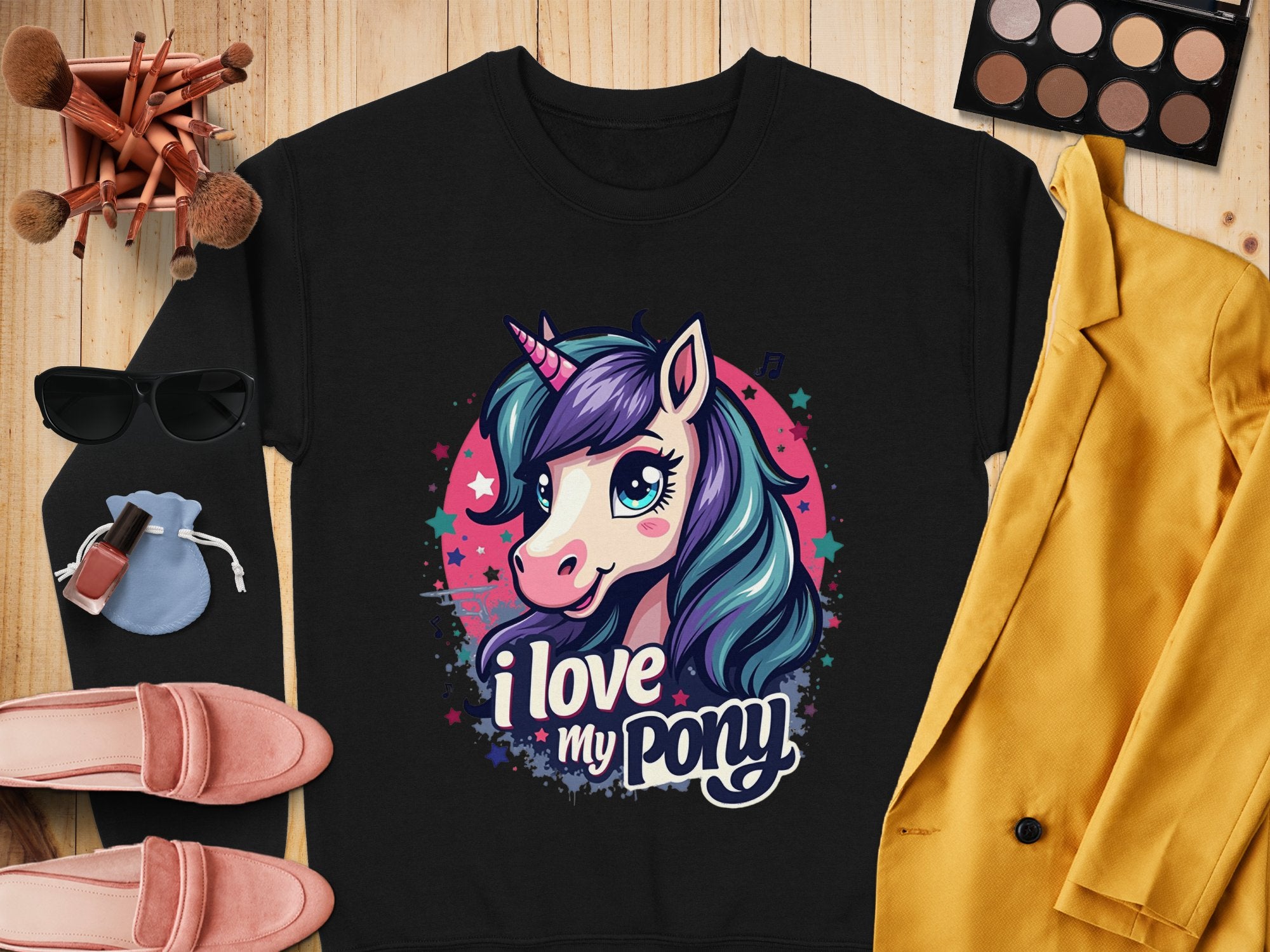 A unicorn enthusiasts perfect tee from Garment Graphics, this black shirt features a vibrant unicorn with colorful hair and horn, surrounded by stars. The text reads I love my pony. Styled on wood with makeup, sunglasses, a yellow jacket, pink shoes, and a small bag.