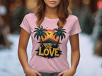 In a blurred outdoor setting featuring snow, a woman wears a Garment Graphics pink T-shirt with two palm trees, a tropical sunset, and a flying plane alongside the phrase One Love, creating warm beach outing vibes.
