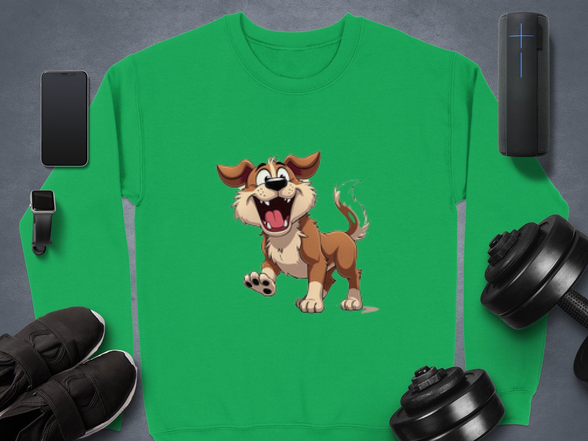 A green Garment Graphics sweatshirt with a happy brown dog illustration and floppy ears, ideal for dog lovers, is displayed on a gray surface surrounded by black sneakers, a smartwatch, a smartphone, a portable speaker, and two dumbbells.