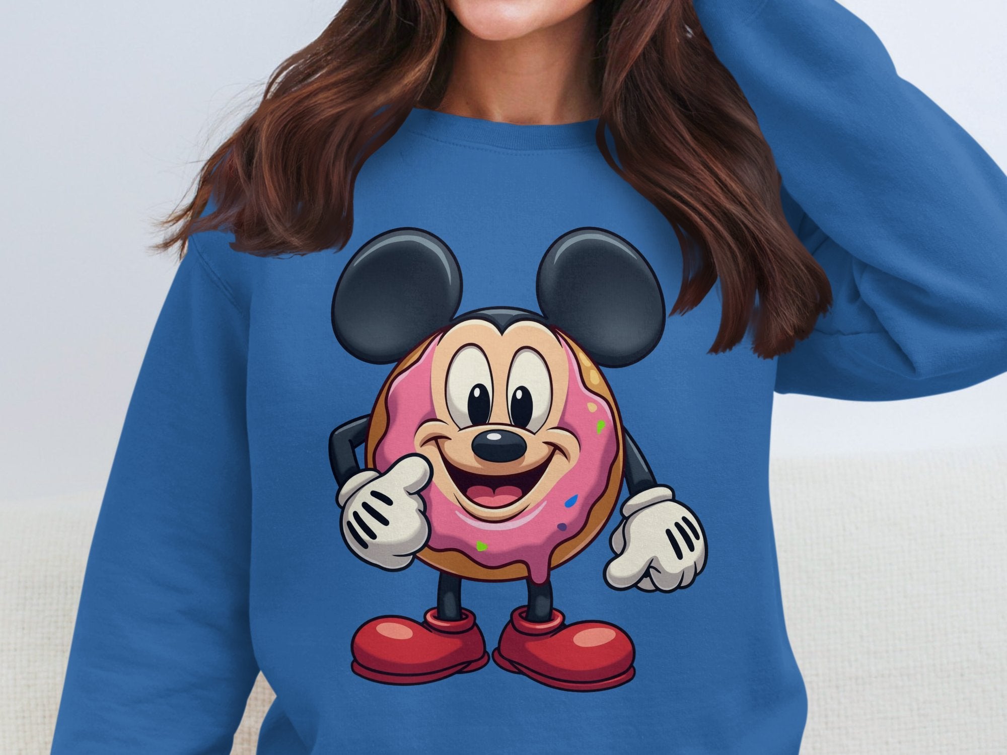 A person wears a unisex blue sweatshirt from Garment Graphics, showcasing a donut character with large ears and a big smile. The playful design features a pink frosted donut with colorful sprinkles, accented by red shoes and gloves.
