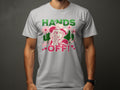 A person wearing a gray Garment Graphics “Hands Off” T-shirt stands against a dark background. The bold design includes a cowboy hat and flowers with vibrant green, pink, and red text. This T-shirt offers a classic fit perfect for casual style enthusiasts.