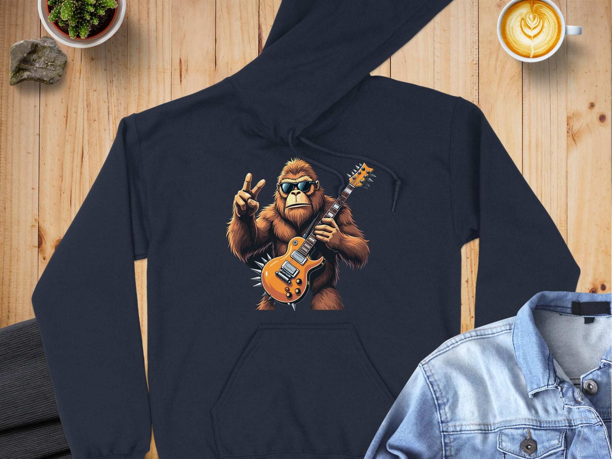 A black hoodie from Garment Graphics showcases a Bigfoot with an electric guitar and peace sign, ideal for music fans. Its set on wood with a denim jacket, potted plant, and latte art coffee.