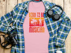 A light pink Born To Be Musician T-shirt by Garment Graphics, ideal for music lovers, showcases drummer and guitarist graphics. Its stylishly placed on a plaid flannel shirt with a camera, wireless headphones, and a takeaway coffee cup on a wooden surface.