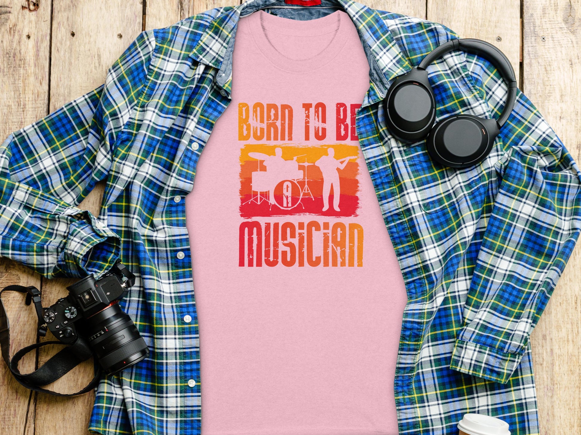 A light pink Born To Be Musician T-shirt by Garment Graphics, ideal for music lovers, showcases drummer and guitarist graphics. Its stylishly placed on a plaid flannel shirt with a camera, wireless headphones, and a takeaway coffee cup on a wooden surface.