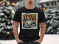 In a snowy setting, an individual wears a classic-fit black T-shirt by Garment Graphics. The shirt features an illustration of a man with the bold text Just Dave, Doing Dave Things.