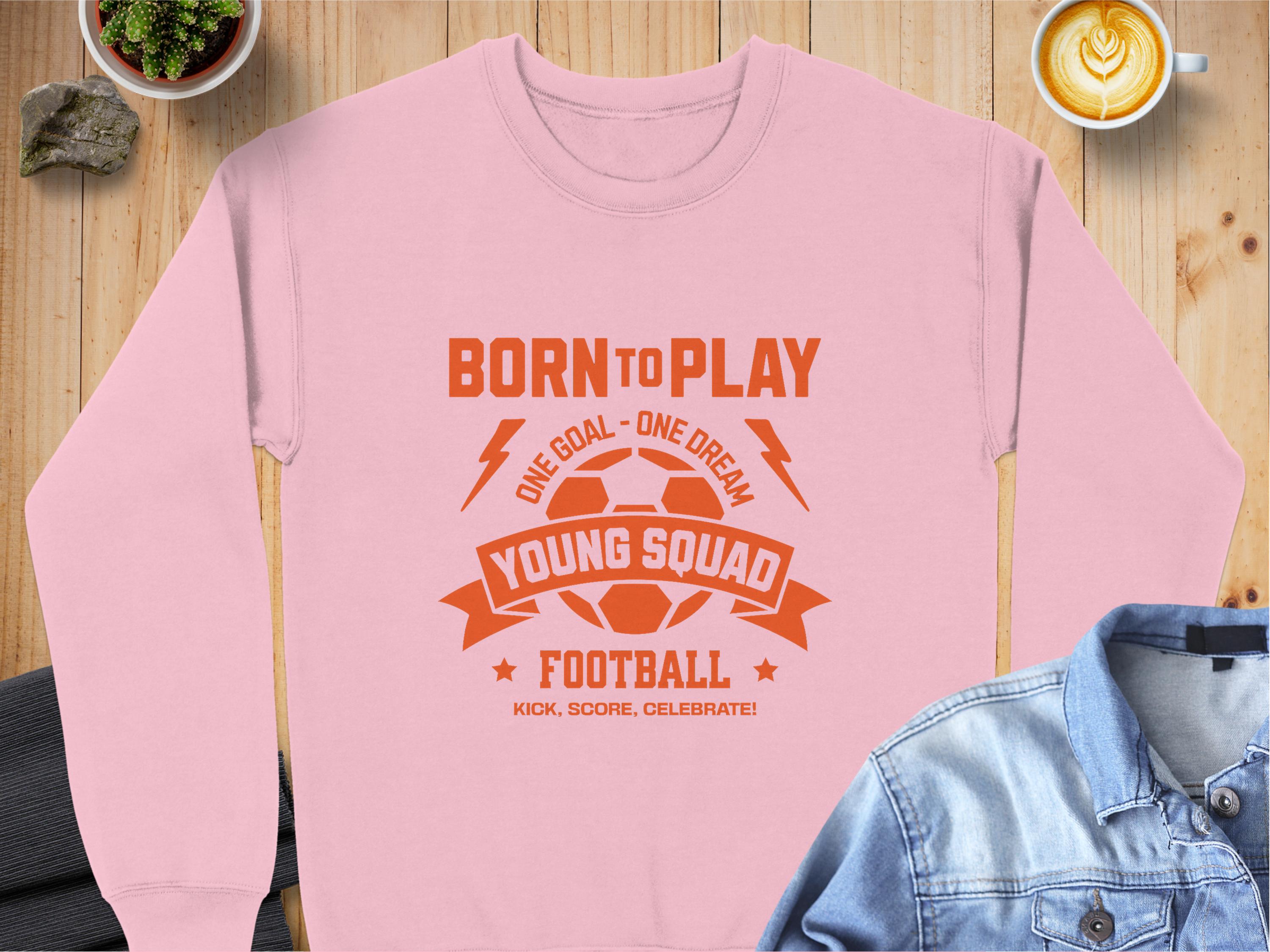 A Garment Graphics pink sweatshirt for young football enthusiasts features an orange graphic with Born to Play, Young Squad, and Football around a soccer ball, plus the phrases One Goal - One Dream and Kick, Score, Celebrate! in unisex sizes on a wooden table.