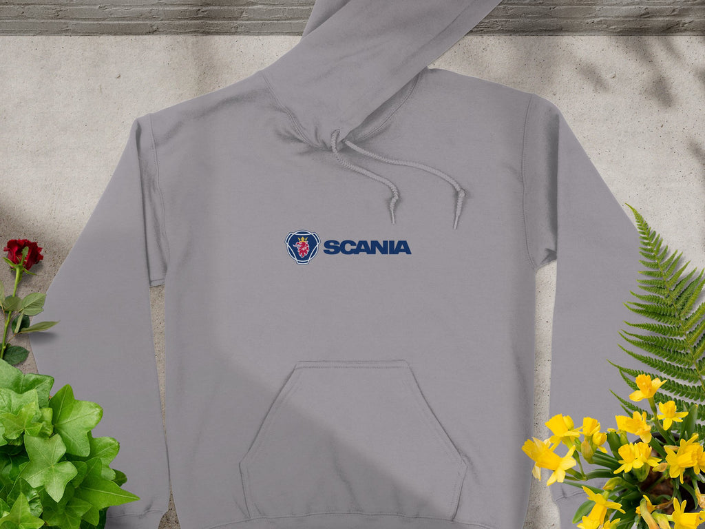 A Garment Graphics gray hoodie with Scania logo and text adorns the chest. This stylish piece, featuring a front pocket and drawstrings, is laid on concrete, surrounded by vibrant green and yellow plants, making it a standout branded hoodie.