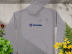 A Garment Graphics gray hoodie with Scania logo and text adorns the chest. This stylish piece, featuring a front pocket and drawstrings, is laid on concrete, surrounded by vibrant green and yellow plants, making it a standout branded hoodie.