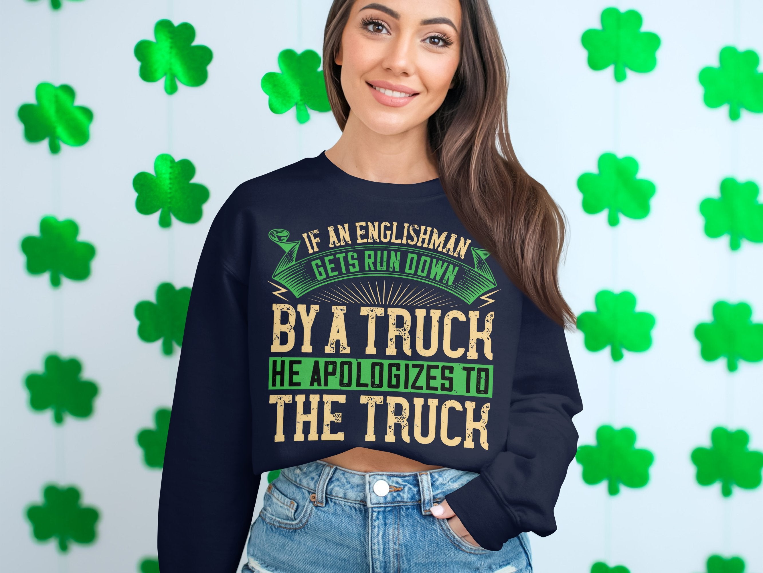 A woman with long brown hair smiles in a navy blue Garment Graphics sweatshirt featuring a funny design: If an Englishman gets run down by a truck, he apologizes to the truck. The unisex sweatshirt is set against green shamrocks.