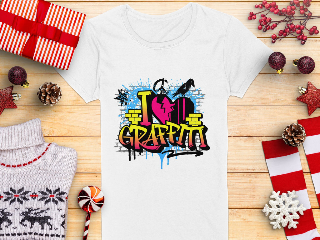 Garment Graphics offers a white t-shirt with vibrant street art, featuring I love Graffiti text with a heart and spray cans. Ideal for urban design lovers, its shown amid wrapped gifts, pinecones, a sweater, a candy cane, and holiday decorations on a wooden surface.