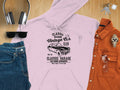 A flat lay from Garment Graphics highlights a pink hoodie featuring a London vintage car graphic, paired with blue jeans. The ensemble is completed with an orange button-up, wristwatch, sunglasses, and headphones on a wooden surface for a classic fit perfect for car enthusiasts.