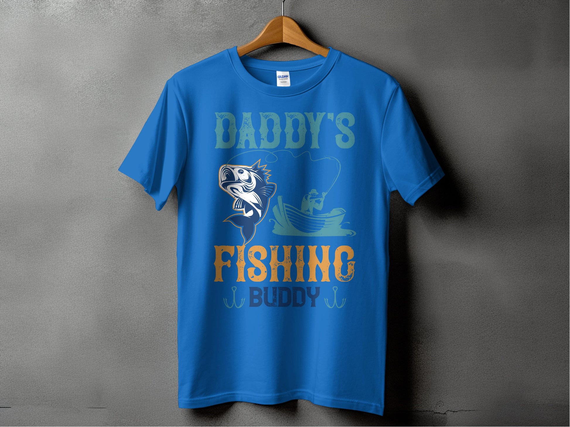 A Garment Graphics blue T-shirt hangs on a wooden hanger against a gray wall. It features a graphic of a fish, person fishing in a boat, and the phrase Daddys Fishing Buddy in vibrant green, blue, and orange, ideal for fishing fans.