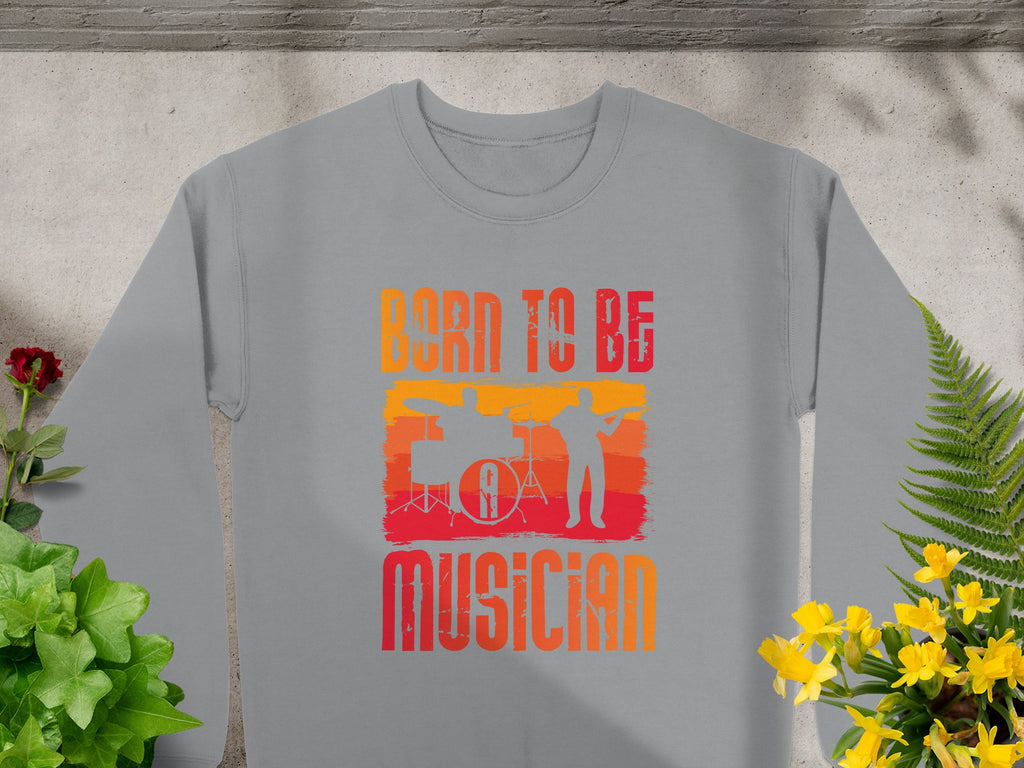Garment Graphics gray sweatshirt, featuring Born to Be Musician text, depicts drummer and guitarist silhouettes on an orange-red background with green plants and yellow flowers. Perfect for music lovers, its available in unisex sizing.
