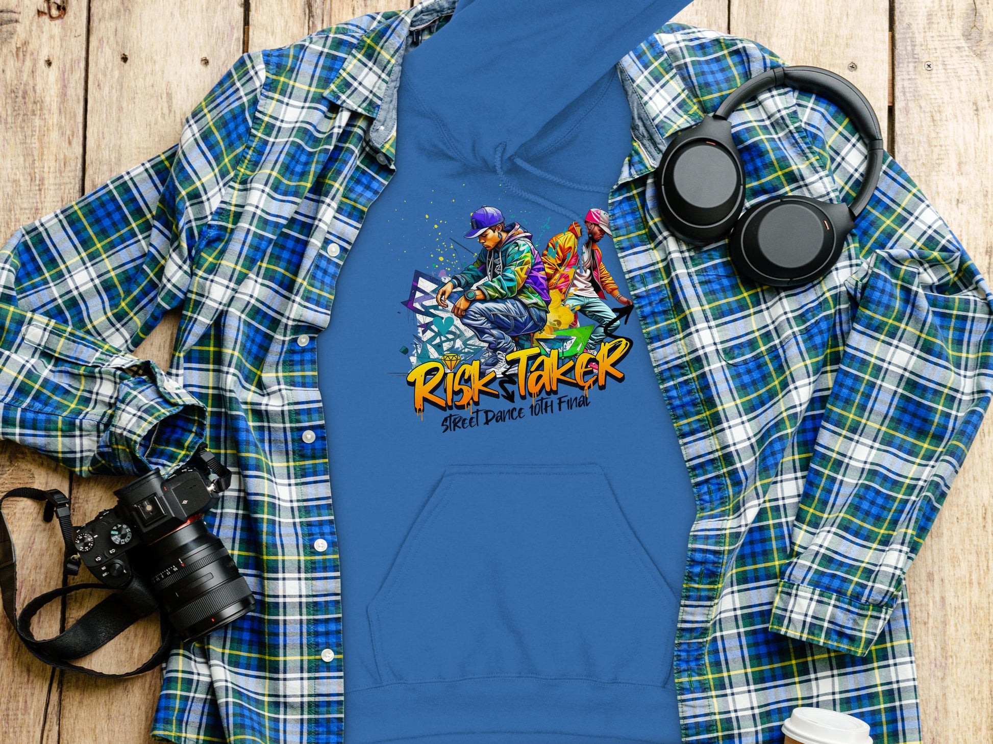 A blue graphic hoodie with Risk Taker, Street Dance with Fire by Garment Graphics is laid on a wooden surface. A plaid shirt, camera, and headphones accompany it, completing the bold fashion statement.