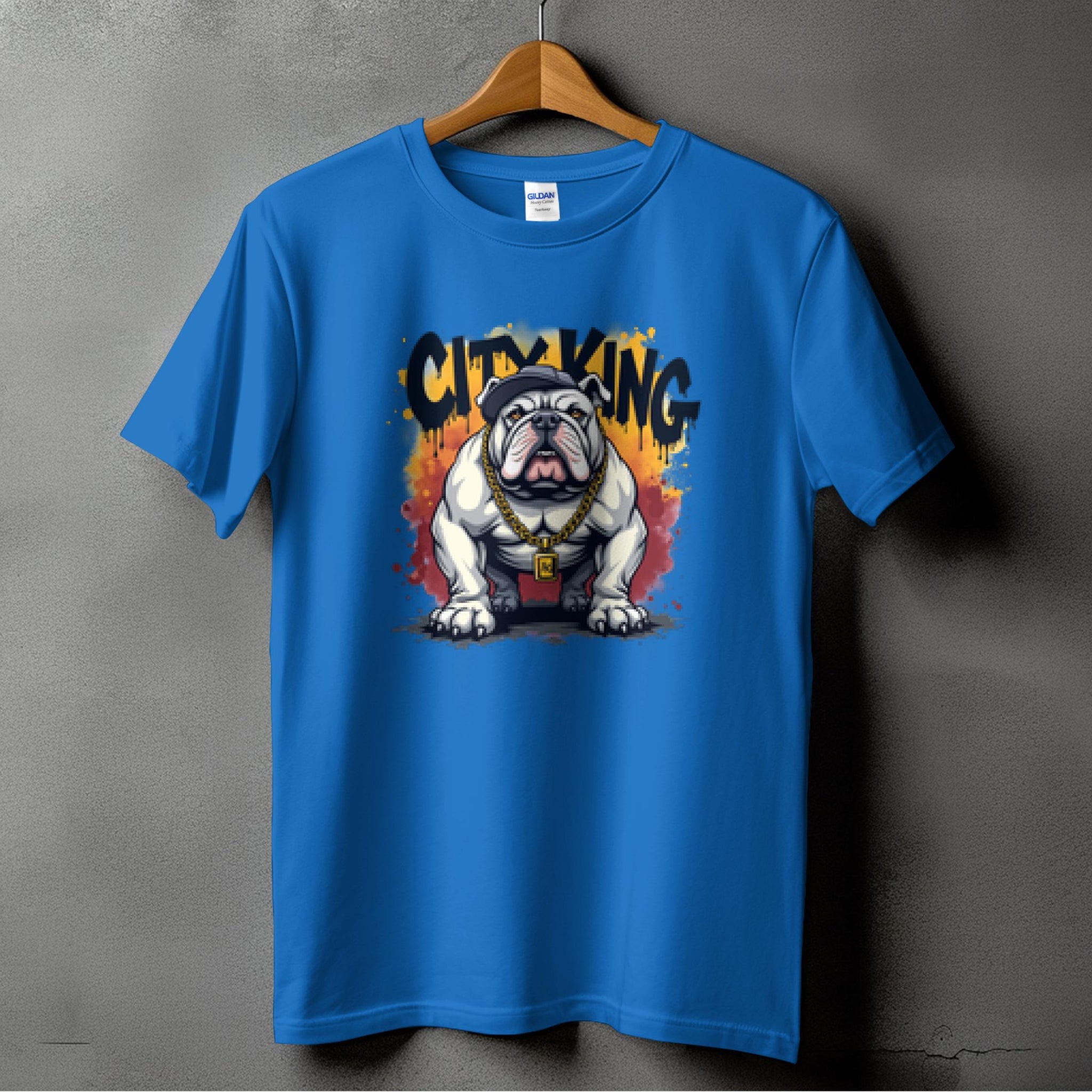 A blue T-shirt from Garment Graphics hangs on a wooden hanger against a textured gray wall. It features a standout bulldog in a red collar with CITY KING and vibrant flames above, exemplifying the unique style of City King T-Shirts.