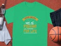 The Garment Graphics green t-shirt featuring Father and Son Fishing Partners for Life with a boat and fish design embodies family bonding, styled on a basketball court next to a basketball, water bottle, black shorts, and gray towel.