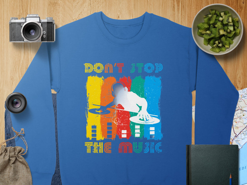 Garment Graphics offers a blue sweatshirt showcasing a DJ design over a colorful abstract background with Dont Stop the Music in playful lettering. Ideal for music enthusiasts, its surrounded by a camera, lens, kiwi bowl, bag, map, and closed book on wood.