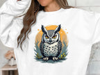 An individual wears a Garment Graphics unisex white sweatshirt featuring an owl with vivid orange eyes, framed by an orange circle and lush foliage, though their face is hidden.