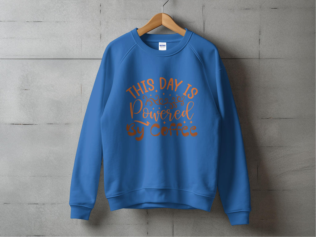 Against a gray concrete wall, a wooden hanger displays a Garment Graphics sweatshirt in blue with a playful design featuring orange text: This Day is Powered by Coffee, adorned with decorative elements.