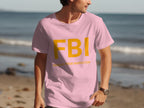 A man wears a light pink Garment Graphics T-shirt on the beach, featuring big yellow FBI letters and Female Boob Inspector beneath. The ocean and sand complement this funny design.